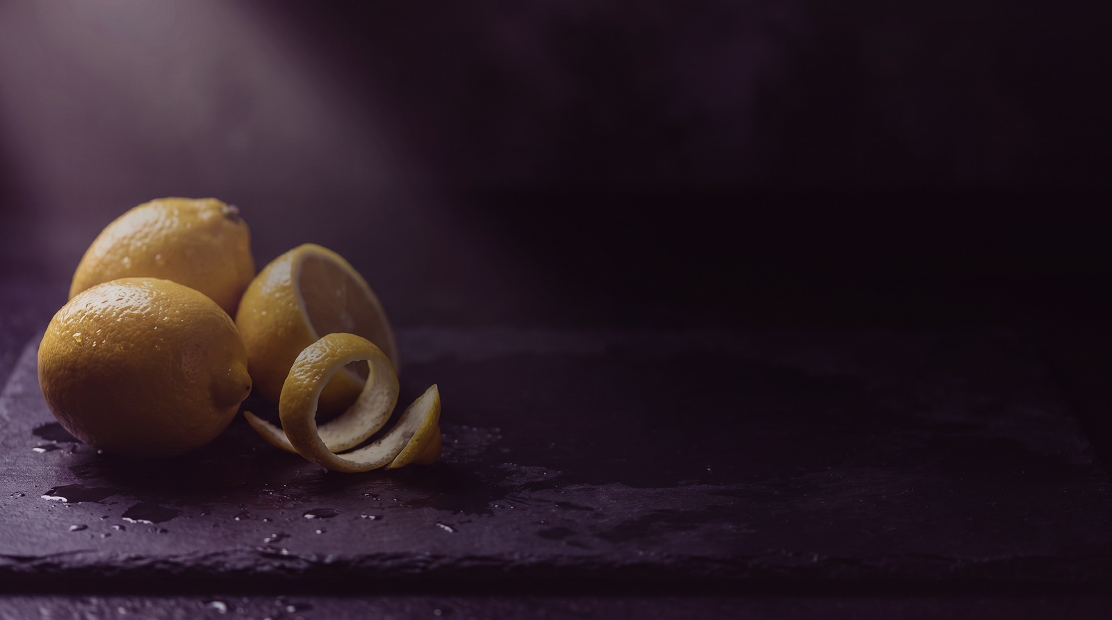Fresh lemons and a curl of lemon peel on a dark wet stone surface, lit from one side in deep violet tones. Header image for an article on the published clinical trials of Eriomin, the standardised lemon flavonoid in Kura Mynis.